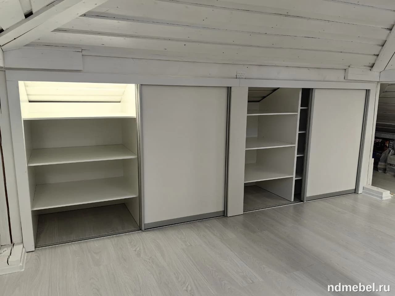 Это встроенный шкаф-купе с раздвижными дверцами, выполненный в минималистичном стиле и окрашенный в белый цвет из материала, похожего на ЛДСП или МДФ. Мебель интегрирована в пространство под наклонным потолком мансарды, что видно по скошенным белым балкам.