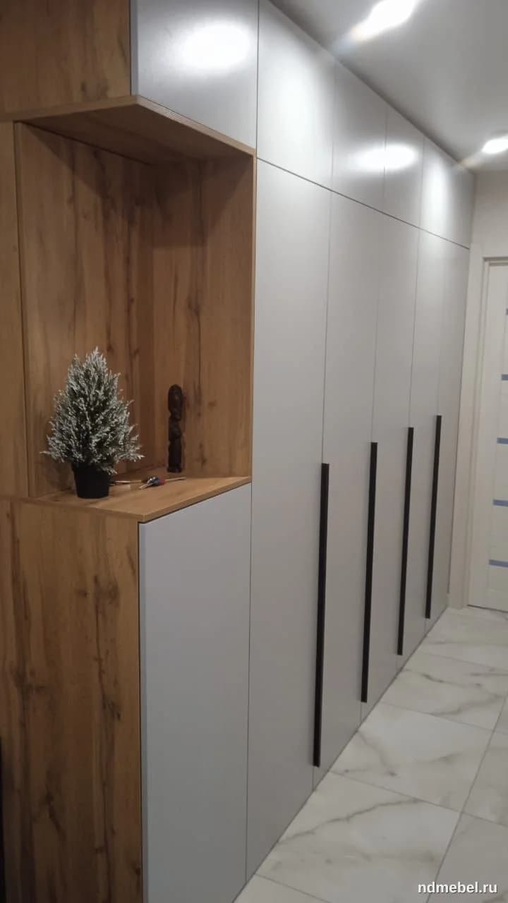 Встроенный шкаф-купе в прихожей, выполненный в современном стиле с комбинацией матового светло-серого и текстурированного дерева (дуб). Мебель имеет минималистичные черные вертикальные ручки и открытую нишу для декора и хранения.