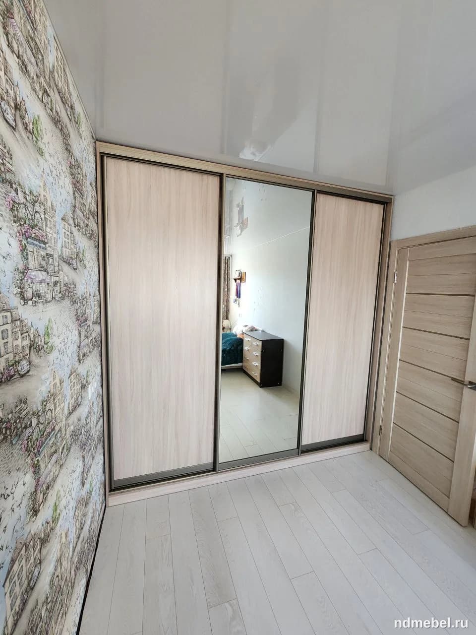 Представлен трехсекционный шкаф-купе с раздвижными дверями, где две секции выполнены под светлое дерево, а центральная — зеркальная. Мебель выполнена в современном стиле и отлично подходит для спальни или прихожей.