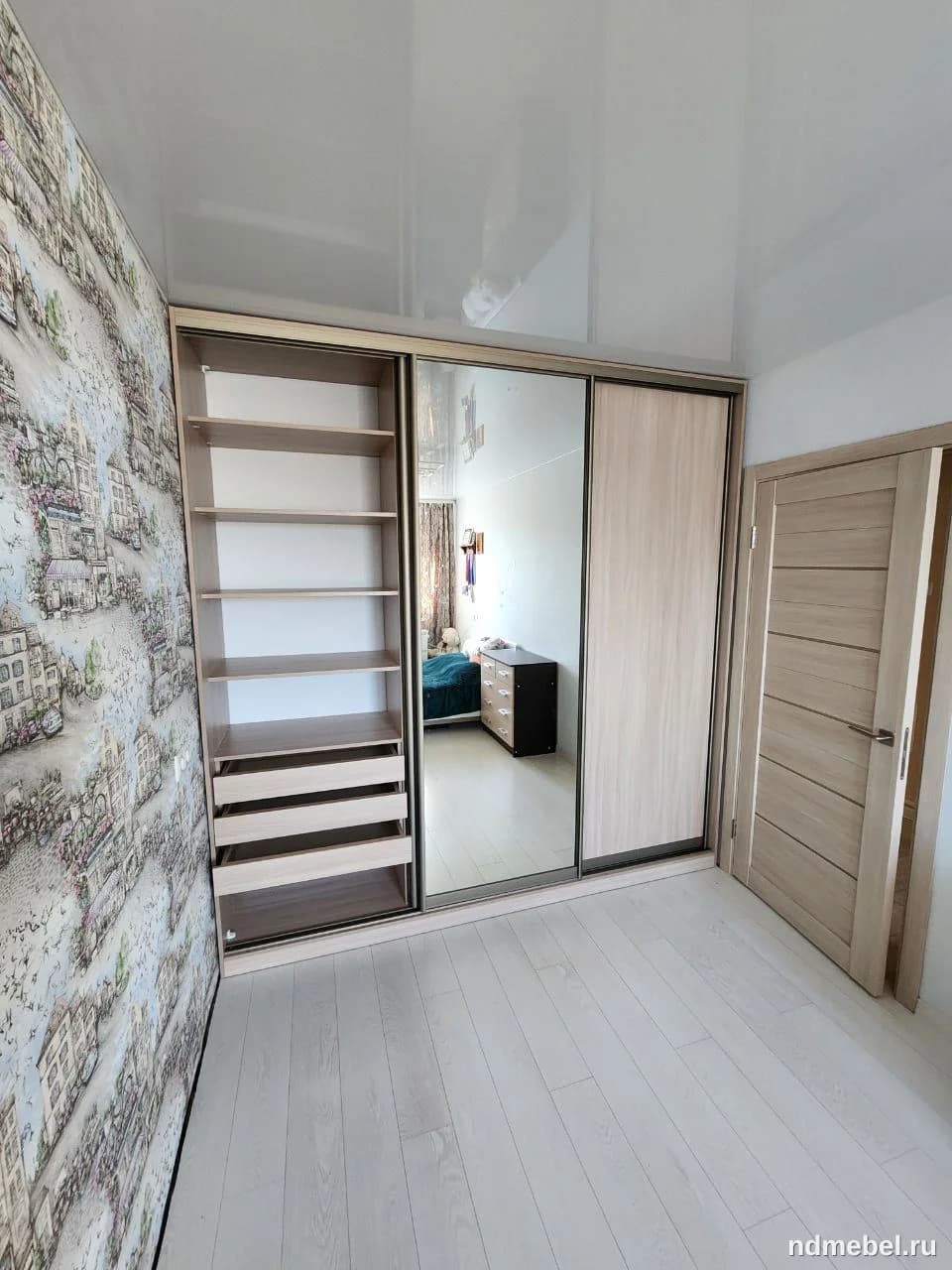 Встроенный шкаф-купе с раздвижными дверями, выполненный в светлом древесном декоре и одной зеркальной вставкой, обладает минималистичным дизайном, дополненным открытыми полками и выдвижными ящиками с одной стороны. Эта мебель прекрасно впишется в современную спальню или гардеробную.