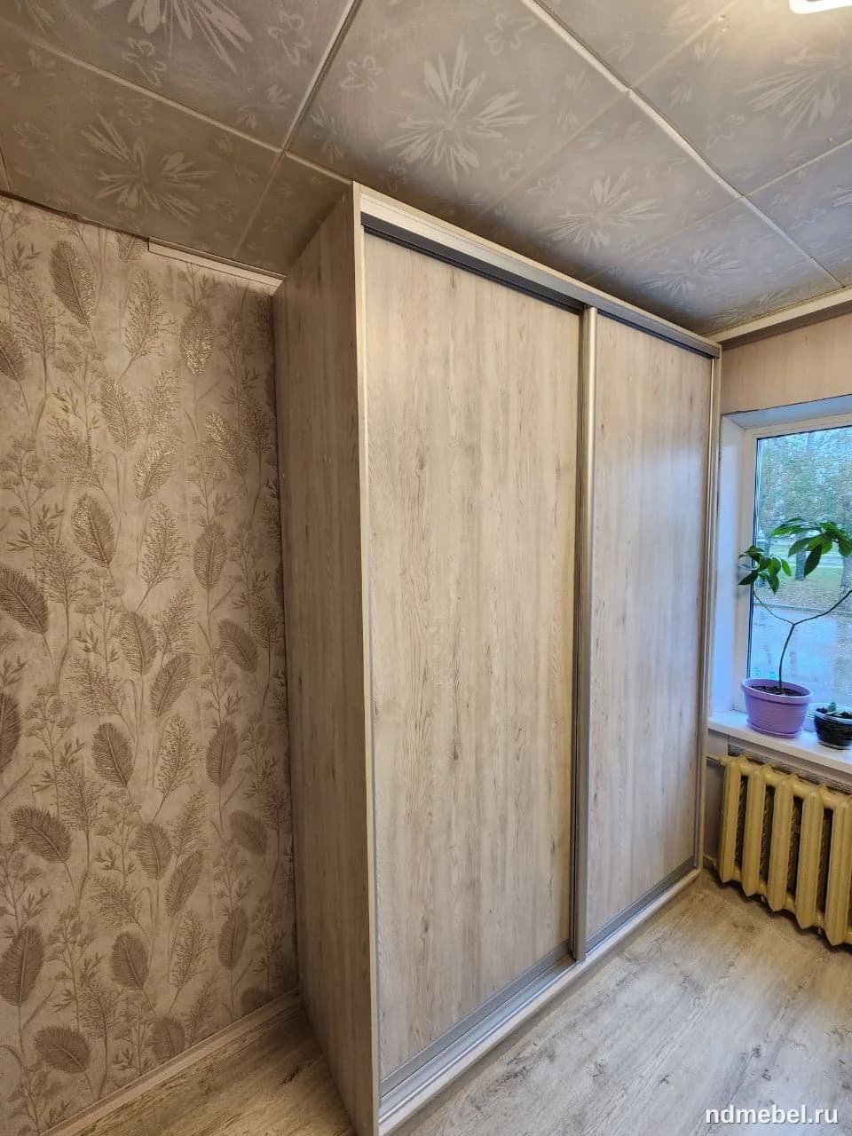На изображении представлен двухдверный шкаф-купе с фасадами под светлое дерево, установленный вплотную к стене. Мебель выполнена в современном, функциональном стиле и оснащена раздвижной системой с серебристыми профилями.