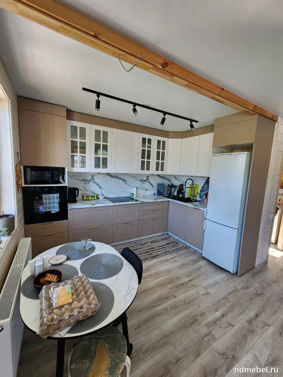 Современная угловая кухня с фасадами двух цветов: бежево-розового снизу и белого со стеклянными вставками сверху, контрастирующими с мраморным фартуком. Пространство дополнено обеденной группой с овальным столом с мраморной столешницей.