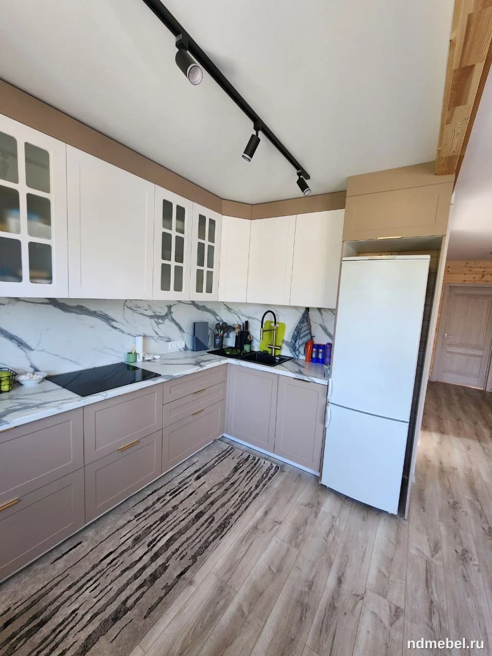 Современный кухонный гарнитур с комбинированными фасадами: нижние секции выполнены в приглушенном бежево-розовом цвете, а верхние — белые (некоторые со стеклянными вставками). Стены рабочей зоны отделаны белым мраморным фартуком, контрастирующим с темной техникой и фурнитурой.