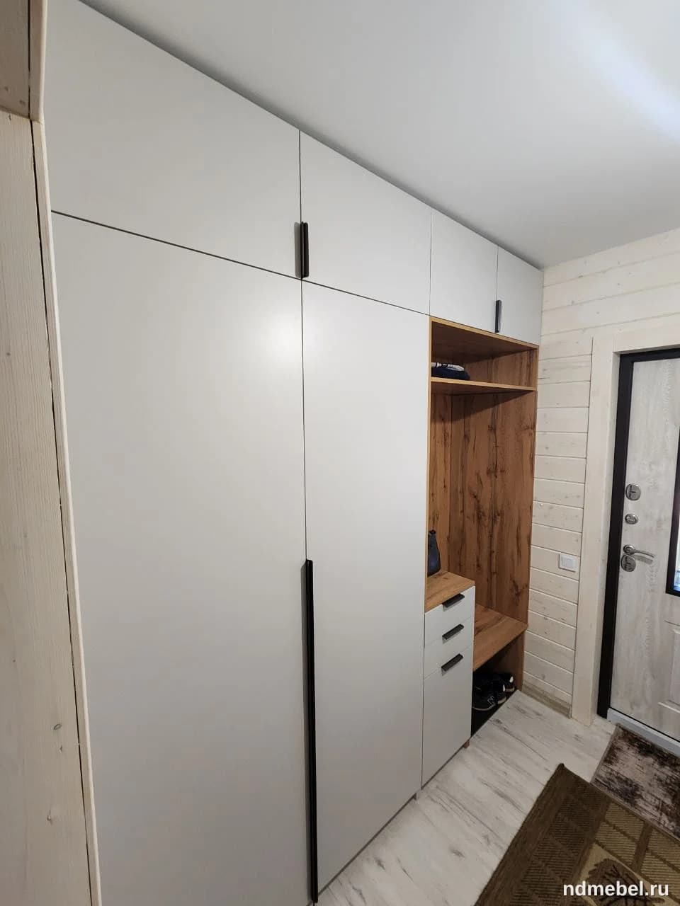 Встроенный шкаф-купе или прихожая система в современном стиле, выполненная в белом матовом цвете с контрастными черными вертикальными ручками и открытыми секциями из шпона или ламината под темное дерево. Мебель занимает всю высоту стены и сочетает глухие фасады с открытым пространством для обуви и мелочей.