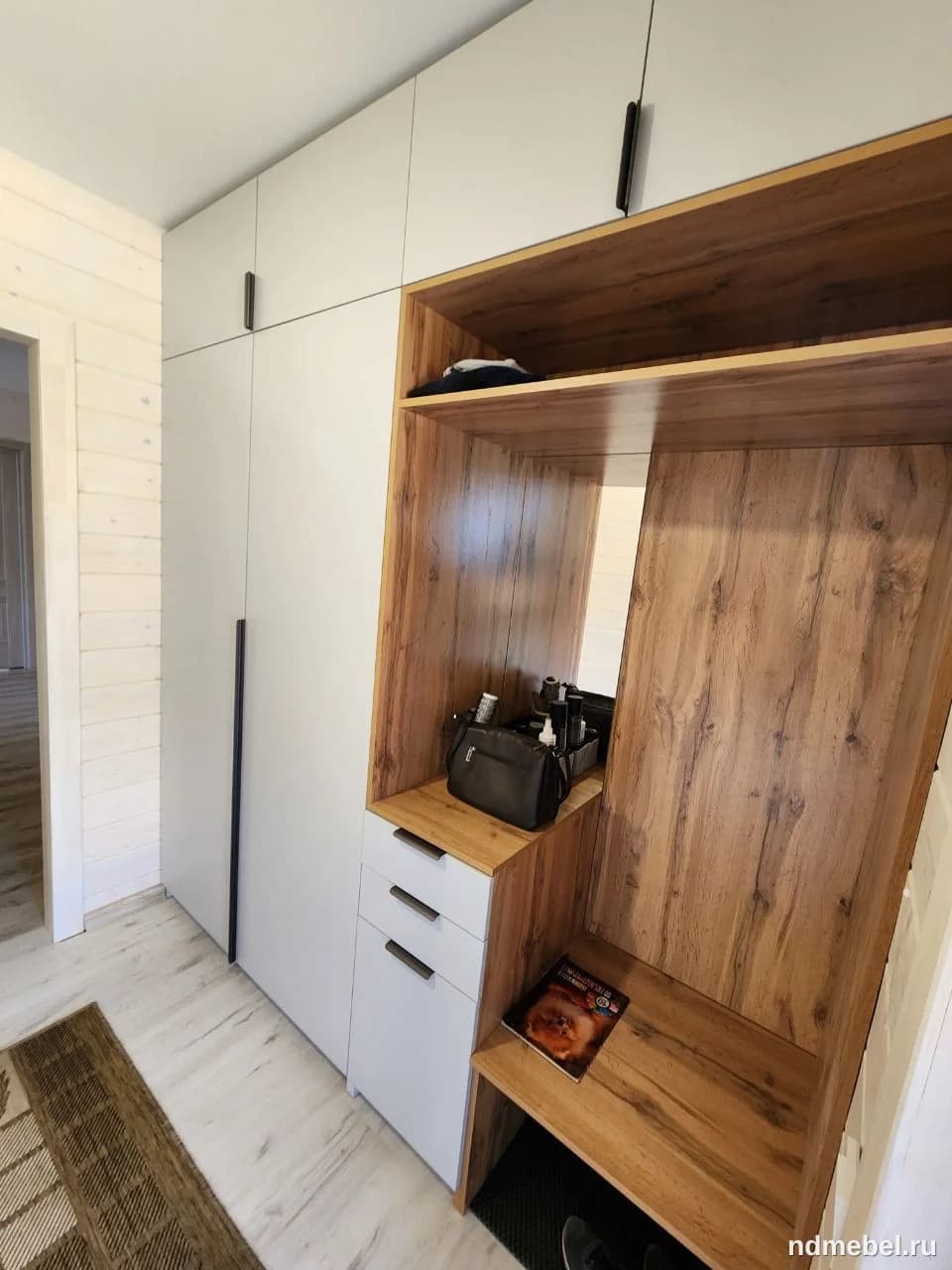 Встроенный шкаф-купе с секцией для прихожей, выполненный в современном стиле, сочетает матовые белые фасады с элементами из ЛДСП под натуральное дерево. Модуль включает закрытые отсеки, открытые полки, зеркало, выдвижные ящики и скамейку для сидения, что характерно для функциональной мебели для коридора.