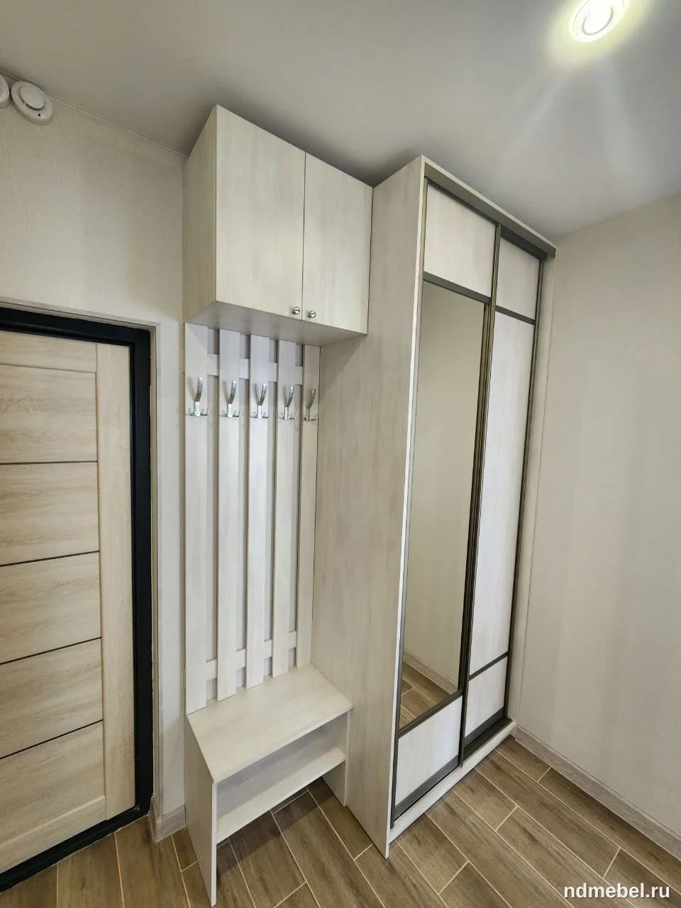 Комплект мебели для прихожей, выполненный в современном стиле и имитирующий светлое дерево или выбеленный дуб. Он включает в себя шкаф-купе с зеркальными дверями, открытую вешалку с полкой-скамьей и навесной шкафчик сверху.