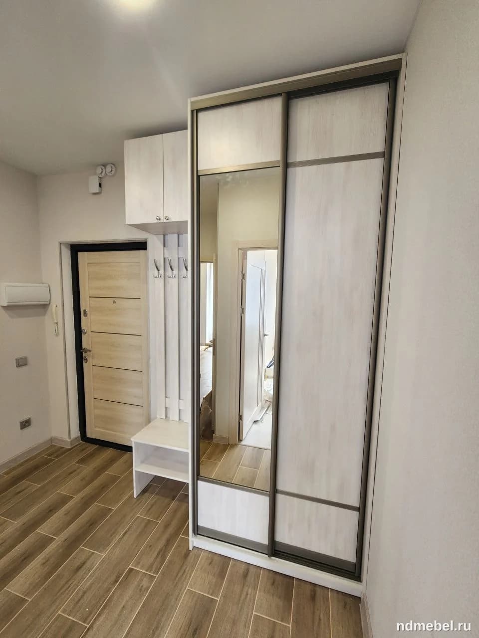 Компактный мебельный гарнитур для прихожей, включающий шкаф-купе с зеркальными дверями и открытую секцию с вешалкой и скамейкой. Мебель выполнена в современном стиле с использованием светлых текстурированных панелей (под светлое дерево) и темной окантовки.