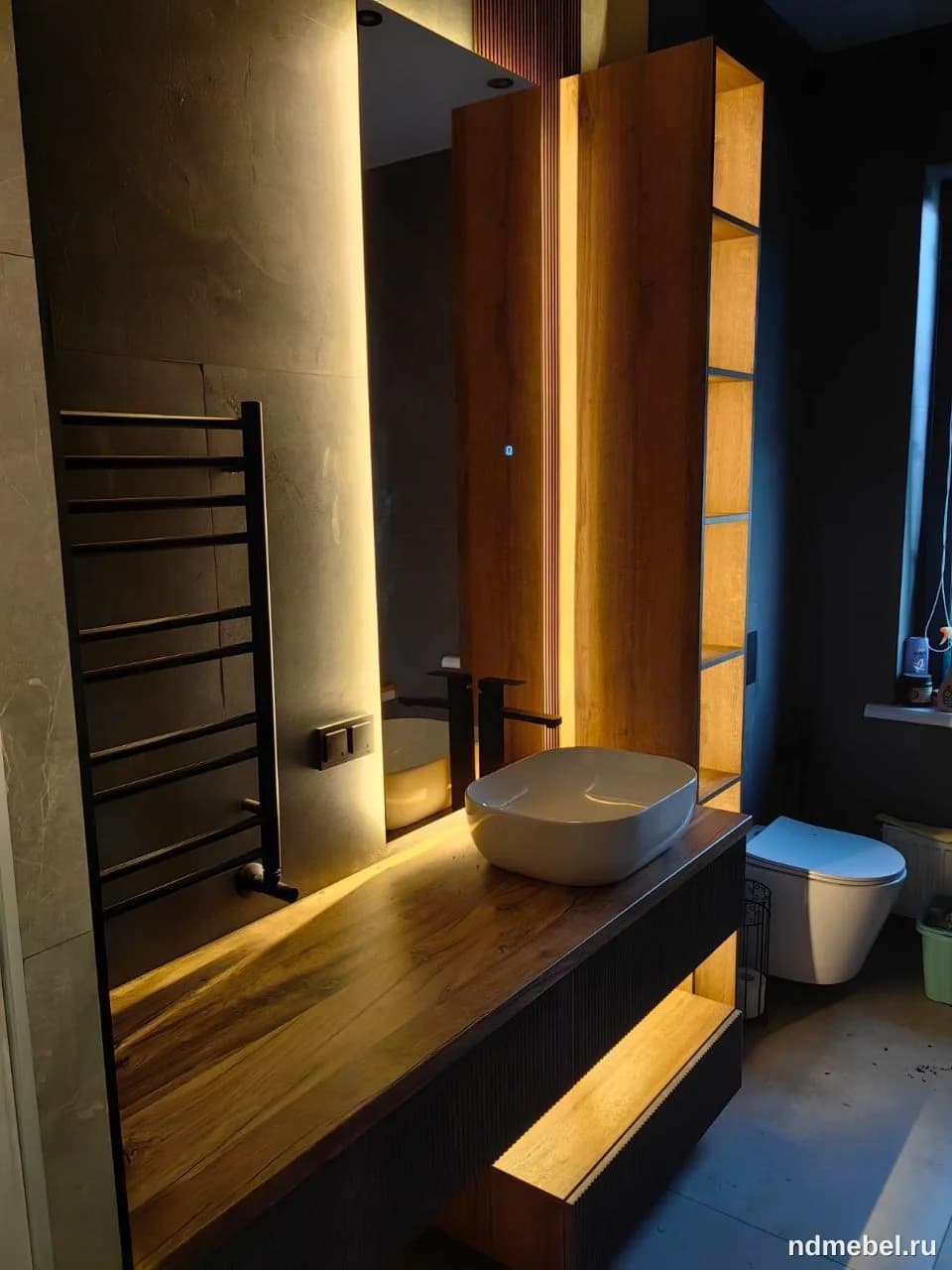 Современная мебель для ванной комнаты, включающая подвесную тумбу с массивной деревянной столешницей и накладной раковиной, а также высокие вертикальные стеллажи с открытыми полками, подсвеченные теплым светом. Мебель выполнена в стиле минимализм с акцентом на натуральное дерево и темную отделку.