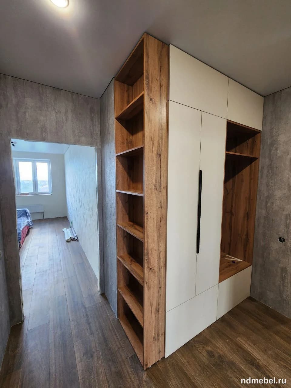 Встроенный угловой шкаф-пенал для прихожей, выполненный в современном стиле с сочетанием светлого матового фасада и декоративных вставок под темное дерево. Мебель включает открытые полки, закрытые секции и нишу для сидения или хранения.