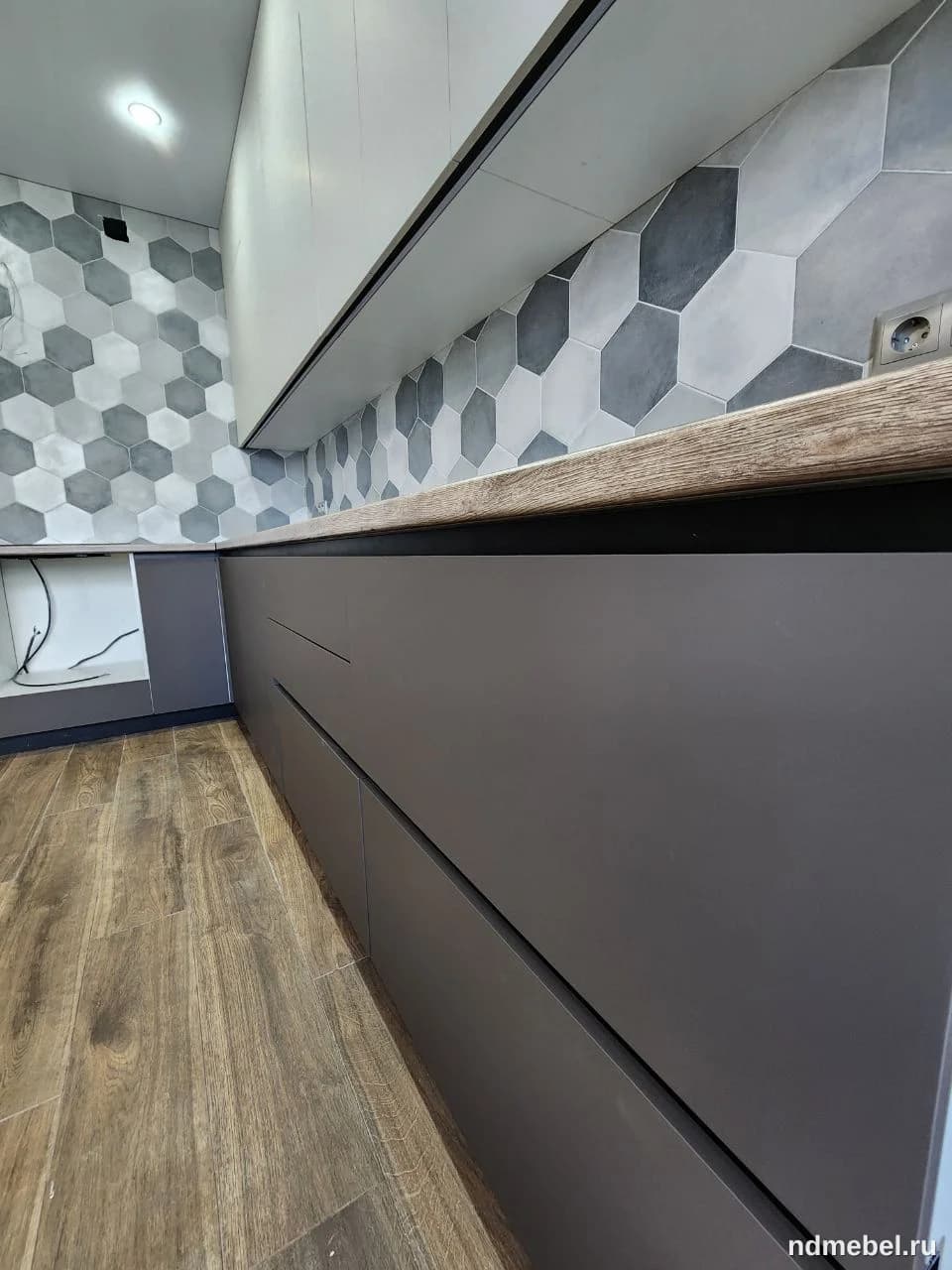 Современная нижняя кухонная линия с гладкими, матовыми фасадами темно-серого (или графитового) цвета и столешницей под натуральное дерево. Дизайн отличается минимализмом с интегрированными ручками или системой push-to-open.