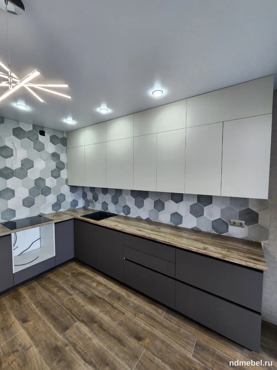 Современный Г-образный кухонный гарнитур с гладкими матовыми фасадами: темно-серые нижние шкафы и белые верхние. Столешница имитирует текстуру светлого дерева, а фартук украшен серой гексагональной плиткой.