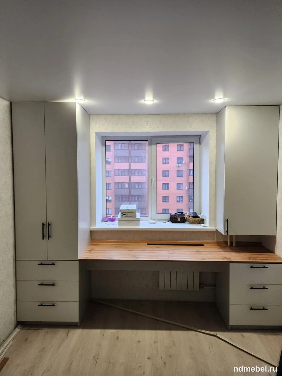 Компактный рабочий комплекс, интегрированный вокруг окна, выполнен в современном стиле с использованием светлого матового материала для корпусов и столешницы под натуральное дерево. Мебель включает боковые пеналы с ящиками и навесными секциями, а также широкую рабочую поверхность, что идеально подходит для организации домашнего офиса или учебного места.
