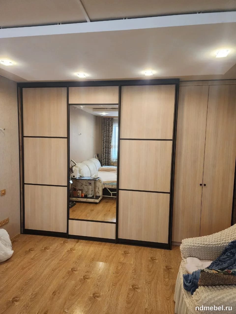 Это большой встроенный или корпусный шкаф-купе с раздвижными дверями, выполненный из светлого дерева (или ЛДСП под дерево) с контрастными темными горизонтальными и вертикальными перегородками. Фасад включает три секции, центральная из которых полностью зеркальная, что характерно для современного или минималистичного стиля.