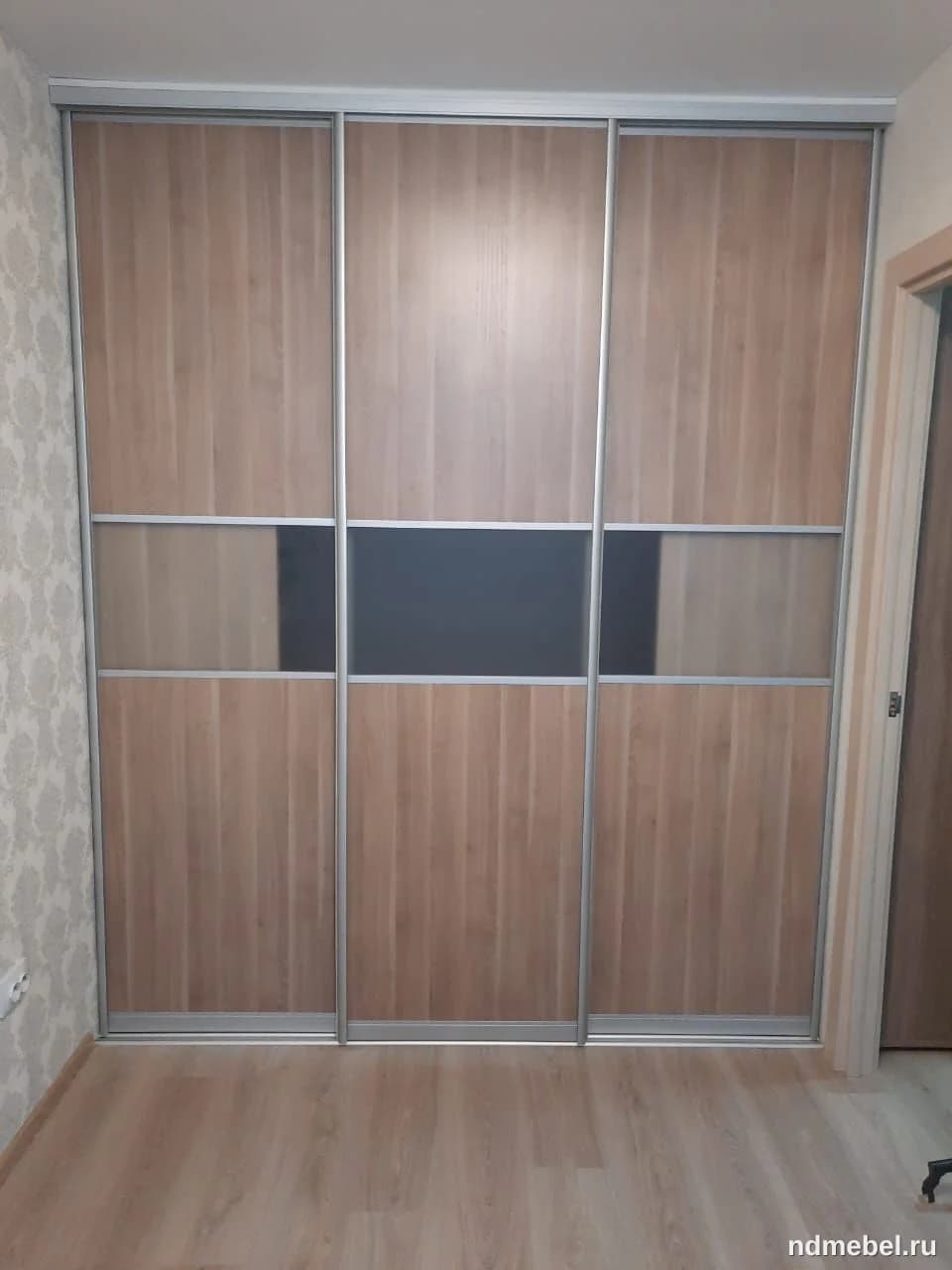 Встроенный шкаф-купе с тремя раздвижными дверями, отделанный панелями под светлое дерево и горизонтальными вставками из матового (или тонированного) стекла серо-коричневого оттенка с алюминиевым профилем. Мебель выполнена в современном стиле и предназначена для хранения одежды.