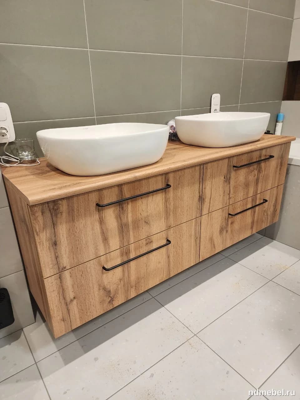 Подвесная тумба для ванной комнаты с имитацией грубого, состаренного дерева дополнена двумя накладными белыми умывальниками и черными матовыми ручками. Мебель выполнена в современном стиле с элементами лофта или скандинавского дизайна.